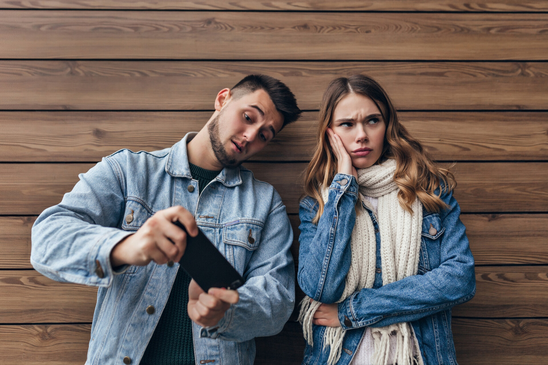 upset charming girl with long scarf looking away while posing wooden wall carefree caucasian man using his smartphone